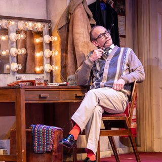 A man with glasses, a striped cardigan, beige trousers and bright orange socks sits in a chair at a dressing table. He holds a pipe in the corner of his mouth and leans one arm on the back of the chair. One leg is crossed over the other.