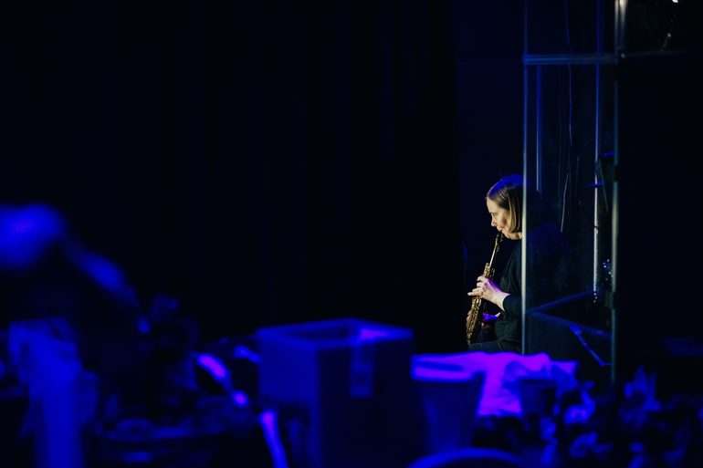 An image taken from backstage. There is a dark curtain covering much of the image with indistinguishable props in the foreground in a very low purple light. A member of the band can just be seen, playing a wind instrument.
