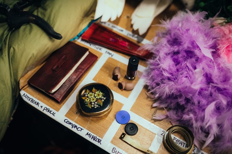 A selection of props arranged neatly on the props table, separated by a grid of masking tape and labelled: a notebook and pencil, a compact mirror, buttons, a fan, feather boas and false severed toes with stage blood on the end.