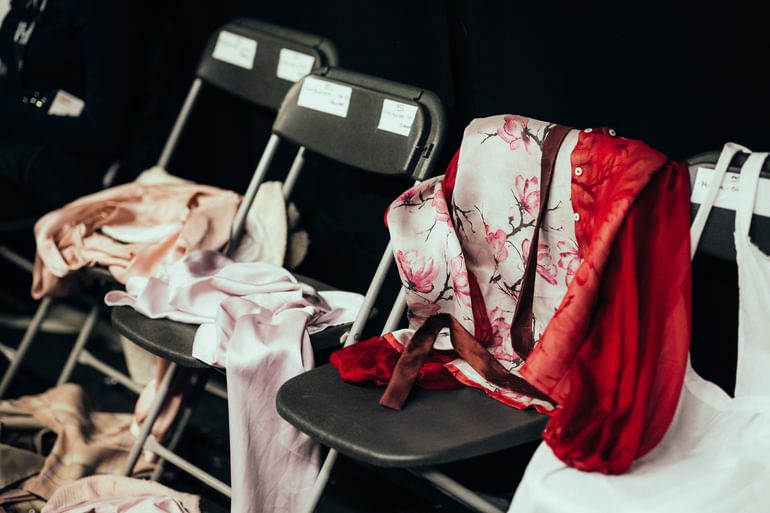 Three grey plastic chairs are lined up with costumes flung over them. There are two pink silk garments and a red silk blouse with a floral pink lining and a burgundy belt.