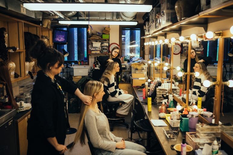 Two performers sit in wigs, hair and makeup having their hair done by two stylists wearing all black. They sit in front of a table packed with hair styling tools and products, in front of a wall of mirrors surrounded with lights. Wigs can be seen on shelves lining the opposite wall.