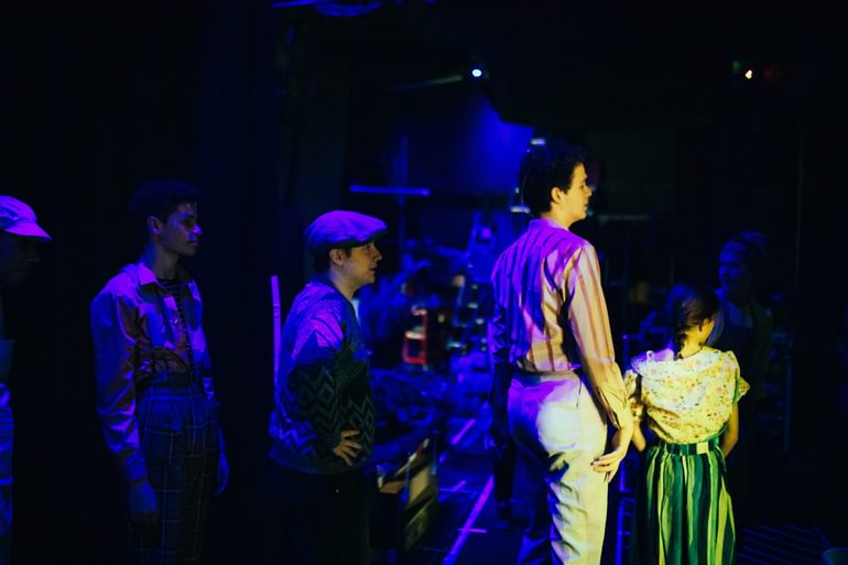 Four performers stand in the dark, dimly lit in blues and purples, waiting to go on stage. They are photographed side on, standing in a line.