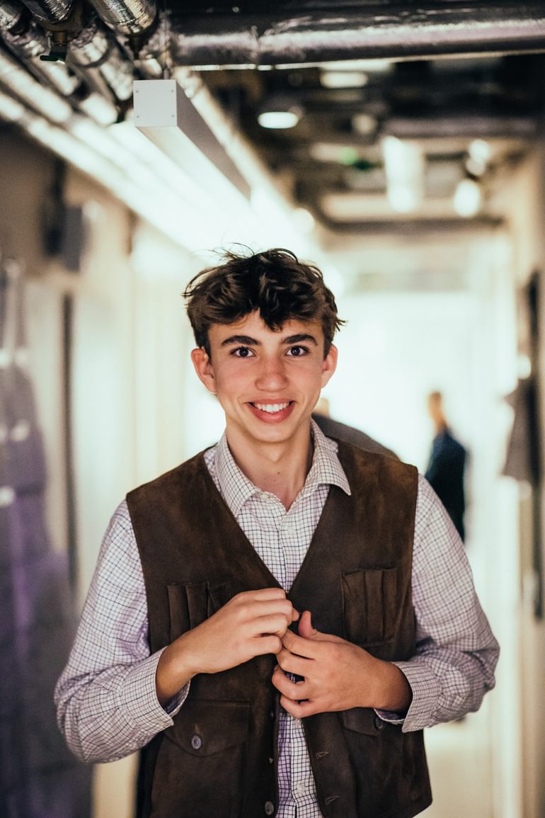 A young performer smiles warmly at the camera. They wear a checked shirt and brown workman's waistcoat, which they are buttoning up.