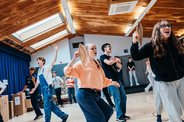 Young performers in casual clothes dance in a rehearsal studio. They are singing and look passionate. Two in the front hold fans and dance with them held up to their faces.