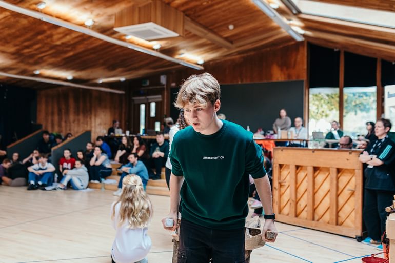 A young performer in casual clothing in a rehearsal studio, with other young performers sitting and looking on in the background, out of focus. They stand dragging a wooden wheelbarrow, singing and looking focused.