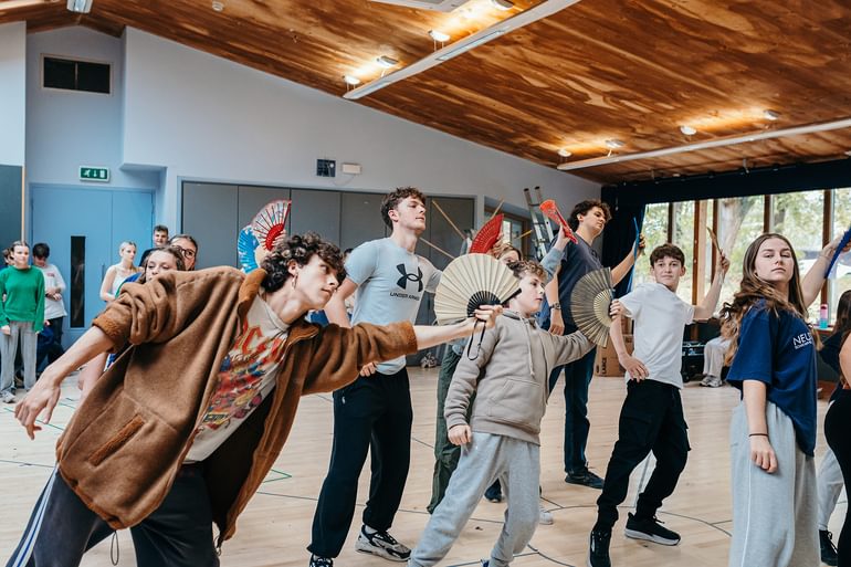 A group of young performers wearing casual clothing dance in a rehearsal studio. They are all holding fans outstretched in their right hands, leaning towards it and looking in the other direction. They look focused and enthusiastic.
