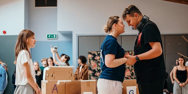 Two young performers clutch hands and stand close together, their foreheads very nearly touching. They look in love. A smaller girl watches on behind them and smiles. In the background other young performers watch, out of focus.