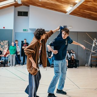 Two young performers stand in a rehearsal room. One holds the other by the neck of his sweatshirt, like he is in trouble. He responds with a shocked facial expression, holding his arms out in desperation. A group of other young performers watch on in the background, out of focus.
