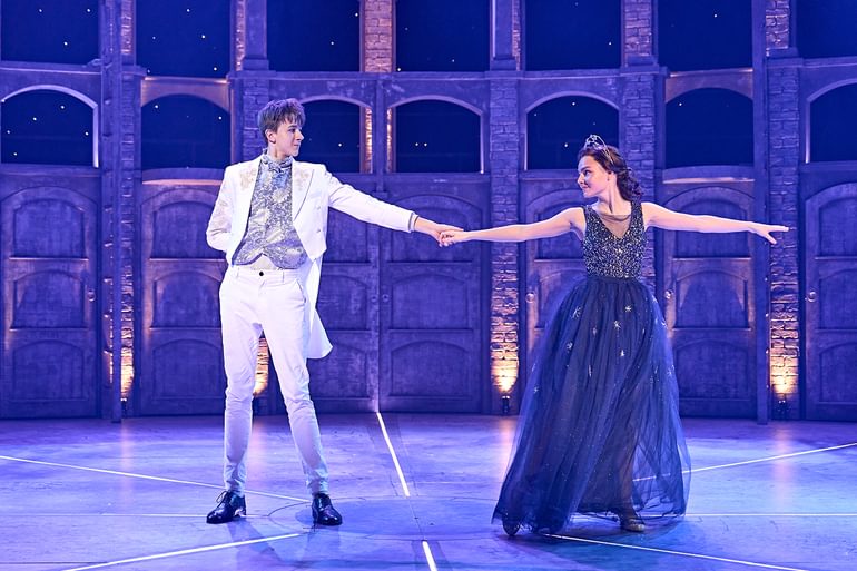 Two young performers dance onstage. They hold their hands in the centre, outstretched and look at one another intently. They look a little shy but happy to be together. The performer on the left wears a white tail suit with a blue floral silk waistcoat and tie. The performer on the right wears a stunning deep blue dress with an embellished bodice and layered tulle skirt scattered with silver flowers with a silver crown in their dark hair, tied back in a bun.
