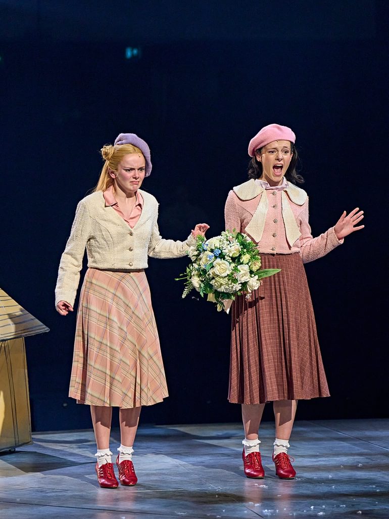 Two young performers stand onstage, looking down with emphatically disgusted facial expressions. They wear coordinated pink outfits with matching red heels and beret hats. One holds a bouquet, that looks like it's from a wedding.