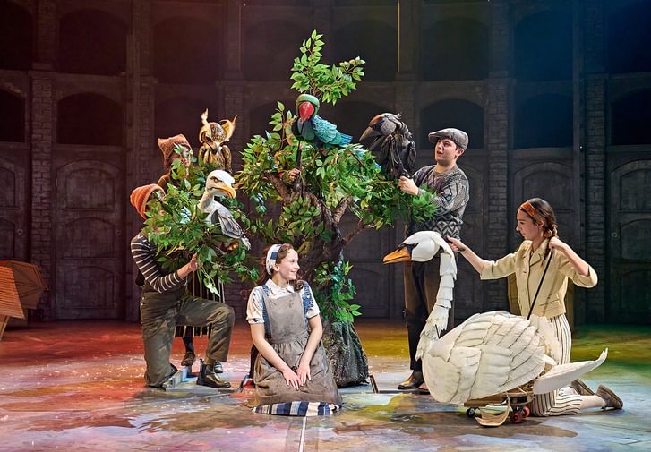 A young performer sits onstage in front of a tree, surrounded by charming, colourful bird puppets: a swan, an owl, a parakeet, a jackdaw and a gull. She looks at the swan with a loving smile. She wears a light brown cotton apron, striped blue and cream skirt, a floral white blouse and a denim waistcoat with hair tied up in a scarf.