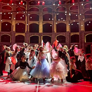 A striking scene onstage with young performers posing in a tableau holding fans, dressed in party costumes. They pose in a variety of different positions and levels, looking up in different directions. The stage is lit in a vibrant red with white light bulbs hanging from the ceiling.