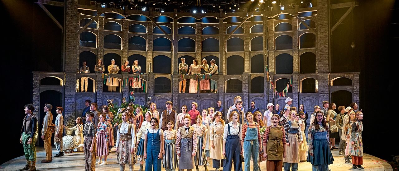 The stage is full of young performers singing enthusiastically. They are all having an amazing time performing. They all wear different costumes like they are from a country village, doing a variety of jobs. They look out to the audience, wide eyed. In the background is a curved wall with lots of windows in, giving the sense of a village behind them.