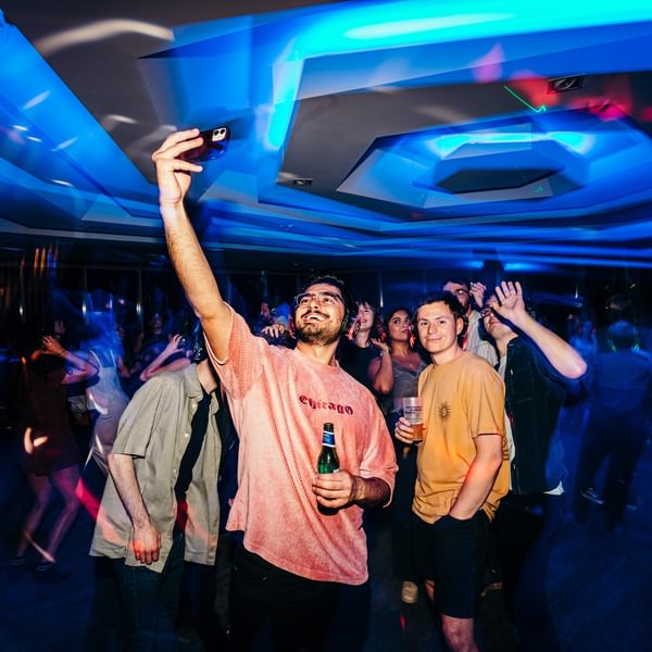 A group of young people pose for a selfie at a silent disco. They are all wearing headphones and smiling. Above them the ceiling is lit blue.