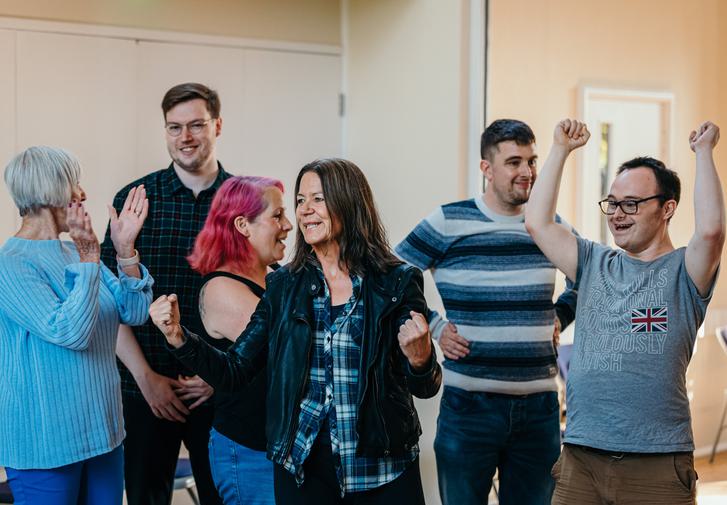 A group of people are all enthusiastically cheering, some are clapping, some clenching their fists and one has their arms raised in the air.