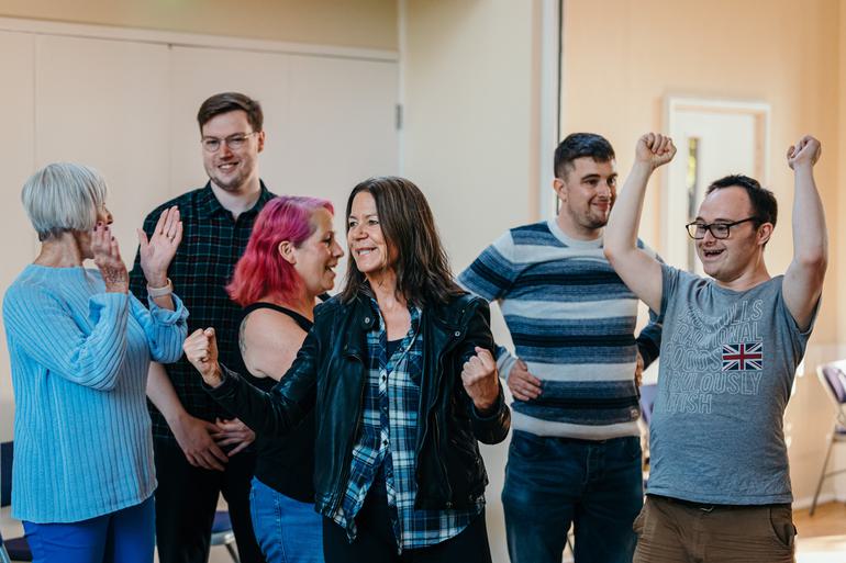 A group of people are all enthusiastically cheering, some are clapping, some clenching their fists and one has their arms raised in the air.