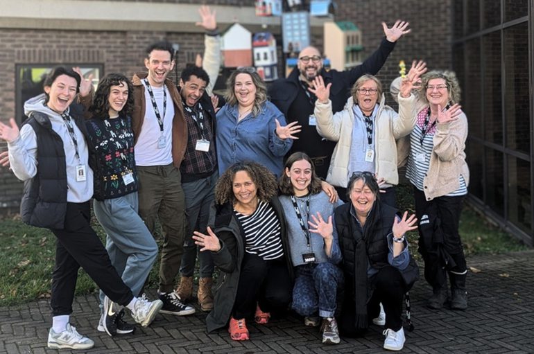 the cast and creative team of Hey! Christmas Tree outside the Minerva Theatre smiling and waving.