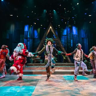 Five performers are hold hands and kicking out in a merry dance. They all wear warm-toned patchwork costumes. Confetti falls from above. Behind them is a wooden a-frame structure and a blue cloth backdrop.