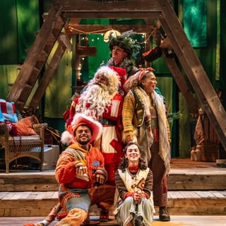 Five performers are gathered in a staggered bunch over three steps. They all wear warm-toned patchwork costumes. They are all smiling, painting a very happy scene. Behind them is a wooden a-frame structure.
