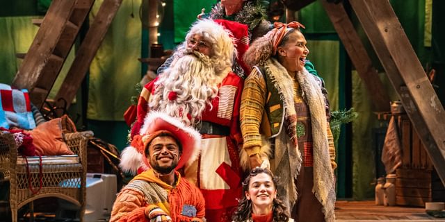 Five performers are gathered in a staggered bunch over three steps. They all wear warm-toned patchwork costumes. They are all smiling, painting a very happy scene. Behind them is a wooden a-frame structure.