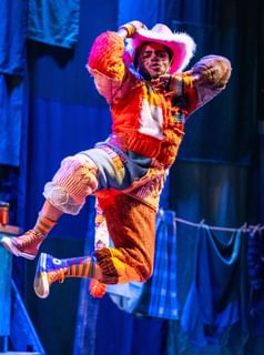 A performer is suspended in mid-air as they click their heels together in a jump. They are dressed as a cat in an orange patchwork outfit, complete with a pink fluffy cowboy hat. They look as if they are enjoying themselves, pulling a pouty expression.