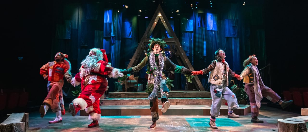 Five performers are hold hands and kicking out in a merry dance. They all wear warm-toned patchwork costumes. Confetti falls from above. Behind them is a wooden a-frame structure and a blue cloth backdrop.