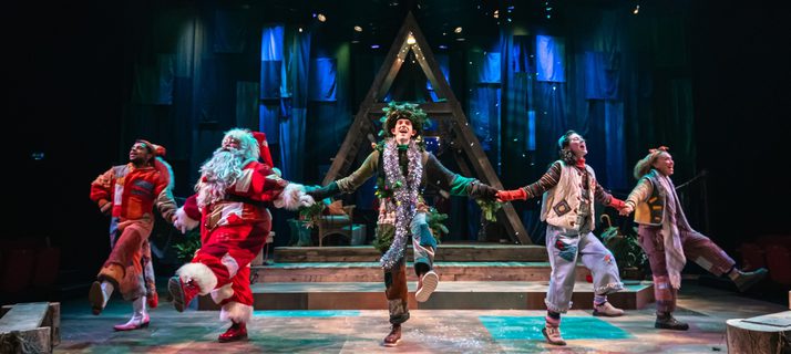 Five performers are hold hands and kicking out in a merry dance. They all wear warm-toned patchwork costumes. Confetti falls from above. Behind them is a wooden a-frame structure and a blue cloth backdrop.