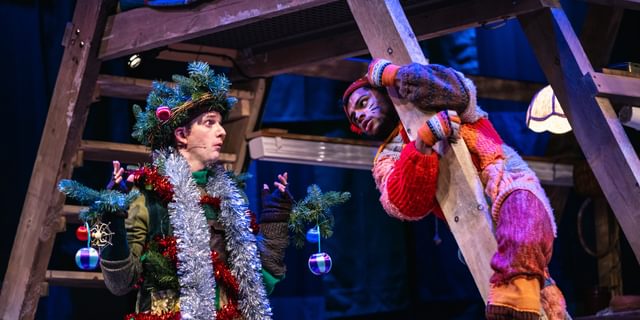 Two performers are talking, the one on the left is dressed as a tinsel covered tree and the other as a cat. Both of their costumes have an earthy, patchwork quality. The performer dressed as a cat is wrapped around a wooden framed structure. The cat looks questioning while the tree looks clueless.