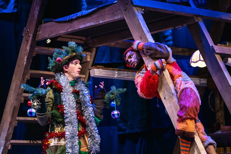 Two performers are talking, the one on the left is dressed as a tinsel covered tree and the other as a cat. Both of their costumes have an earthy, patchwork quality. The performer dressed as a cat is wrapped around a wooden framed structure. The cat looks questioning while the tree looks clueless.