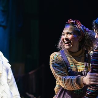 A woman holds the arm of the young girl next to her, she is beaming with a joyful smile. They are dressed in earthy toned, patchwork clothes. Behind them, another performer is dressed as a snowman, complete with top hat.