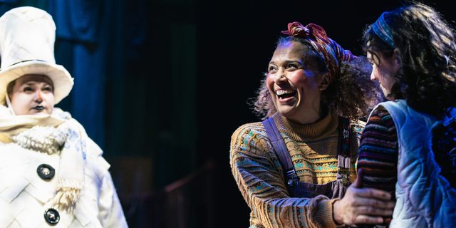 A woman holds the arm of the young girl next to her, she is beaming with a joyful smile. They are dressed in earthy toned, patchwork clothes. Behind them, another performer is dressed as a snowman, complete with top hat.