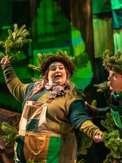 Two performers are dressed as Christmas trees in green patchwork overalls. They wear pine wreaths on their heads and hold branches in their hands. Behind them is a patchwork green cloth backdrop. Their expressions are apprehensive.