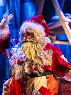 A performer dressed as Father Christmas in a red and white patchwork costume is waving. They wear a big white beard and white gloves. In the background there’s a wooden framed structure in a wash of blue light.