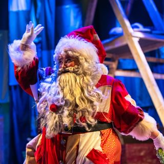 A performer dressed as Father Christmas in a red and white patchwork costume is waving. They wear a big white beard and white gloves. In the background there’s a wooden framed structure in a wash of blue light.