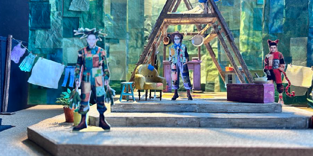 A set model of a green triangular shaped house with washing hanging to the left; three illustrations of characters placed around the stage.