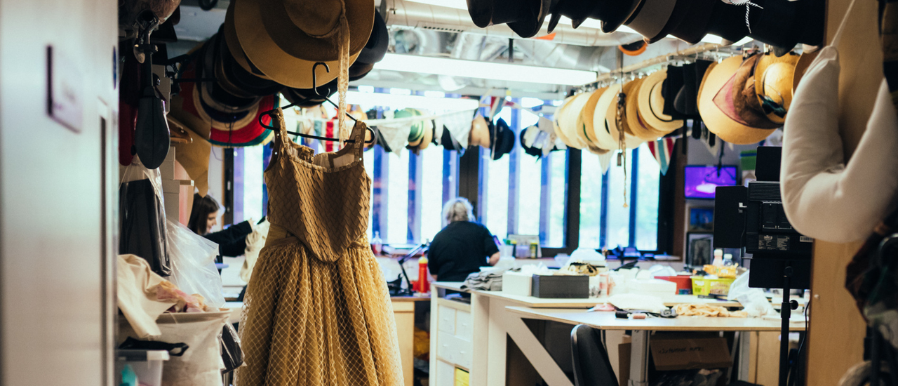 Image taken from the perspective of someone standing in the doorway of our wardrobe workshop, hats of all varieties hang from the ceiling and a dress is waiting to be steamed.