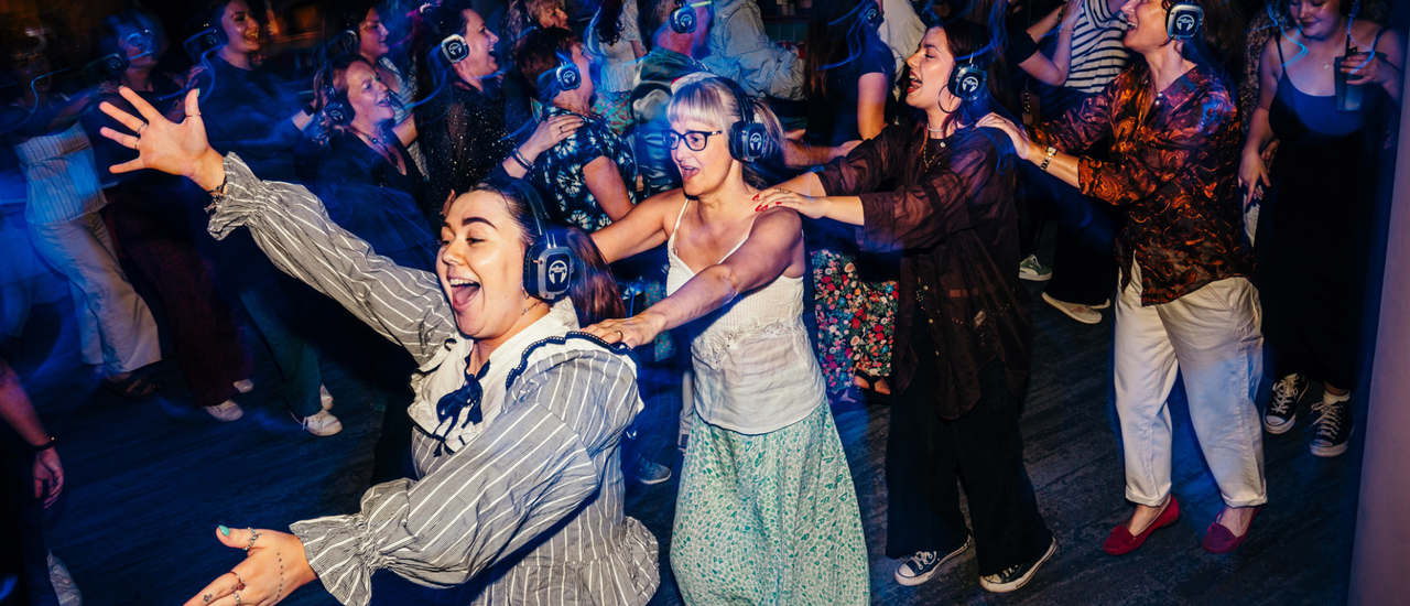 An image of a conga line at a silent disco. Everyone is wearing headphones and laughing with joy.