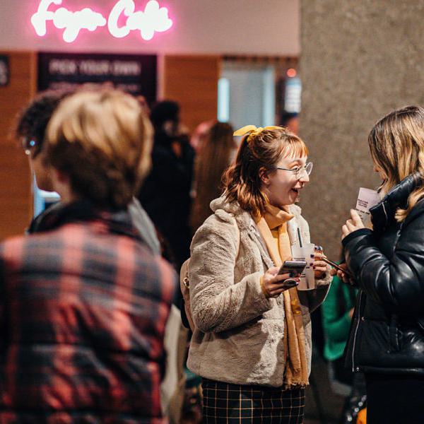 Two women stand in the Festival Theatre foyer, smiling at eachother. They are holding drinks and phones. In front of them are two other people with their faces obscured. In the background is a glowing pink sign which says 'Foyer Cafe'