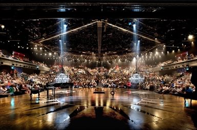The Festival Theatre auditorium is seen from the stage. The audience is full. The black and gold stage stretches out in front of the camera. On the left of the frame is a grand piano. Two white crystal chandeliers hang down from the ceiling. There are lots of golden lights shining towards the stage and the camera from the ceiling