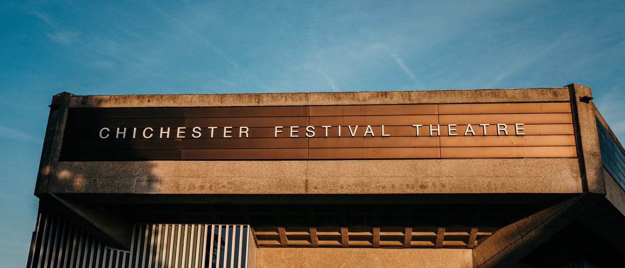 We're looking up at the listed Festival Theatre building, a brutalist design, against a clear blue summer's evening sky. The theatre appears to be lit by late afternoon sun and appears almost gold in colour.