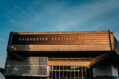 We're looking up at the listed Festival Theatre building, a brutalist design, against a clear blue summer's evening sky. The theatre appears to be lit by late afternoon sun and appears almost gold in colour.