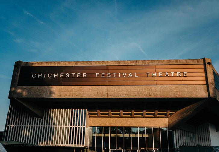 We're looking up at the listed Festival Theatre building, a brutalist design, against a clear blue summer's evening sky. The theatre appears to be lit by late afternoon sun and appears almost gold in colour.