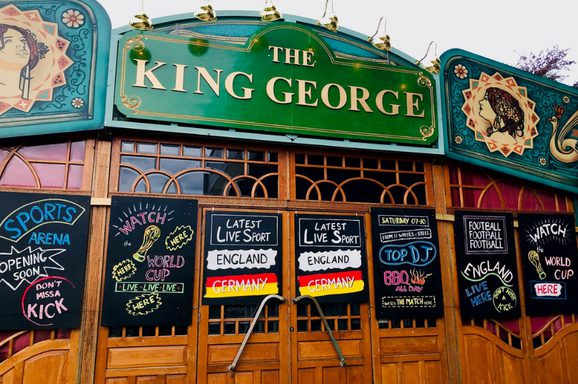 Front view of a theatre-style building with a large green sign reading ‘The King George,’ wooden doors below, and colorful chalkboards advertising live sports and events.