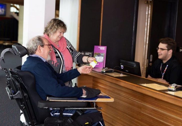 A woman and a man in a wheelchair is speaking with a Box Office assistant.
