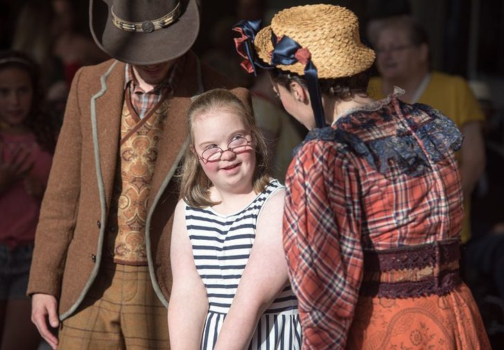 A girl meeting two cast members at a performance, one is dressed in a brown blazer, tweed trousers and cowboy hat, the other in an orange dress and straw hat.