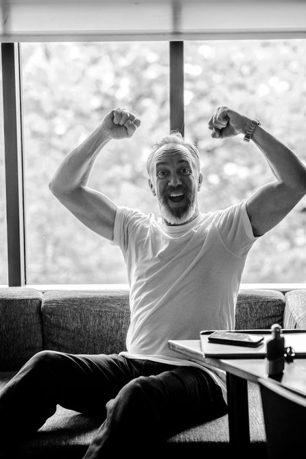 A black and white phot of an actor wearing a white top and jeans, smiling brightly and holding his arms in the air in a celebratory way.