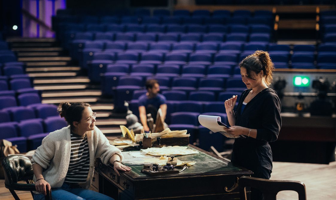 Two women are on stage one sat at a large old-fashioned desk, and the other standing holding a clipboard, they are both smiling at each other.