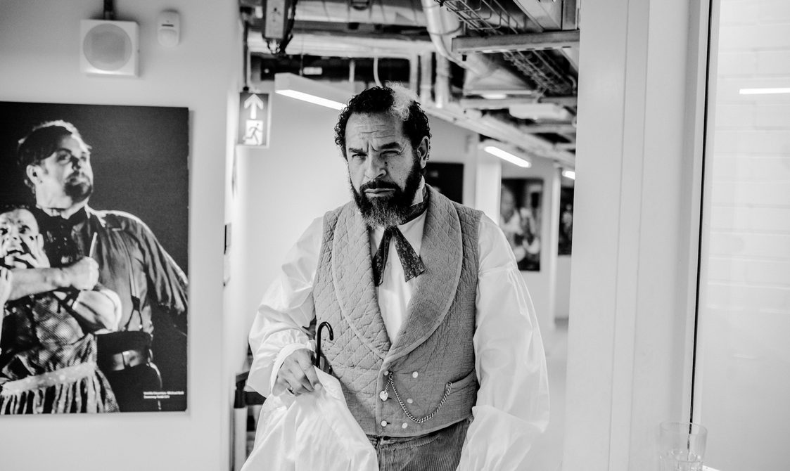 A black and white image of an actor backstage he is wearing his costume and carrying a shirt on a hanger, he is pulling a serious face but there is softness and silliness to it.