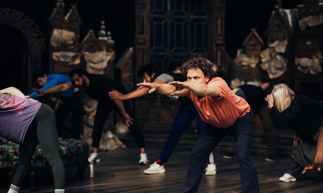 A photo taken on stage during warm-up, in the foreground we see an actor squatting and reaching forward looking very serious.