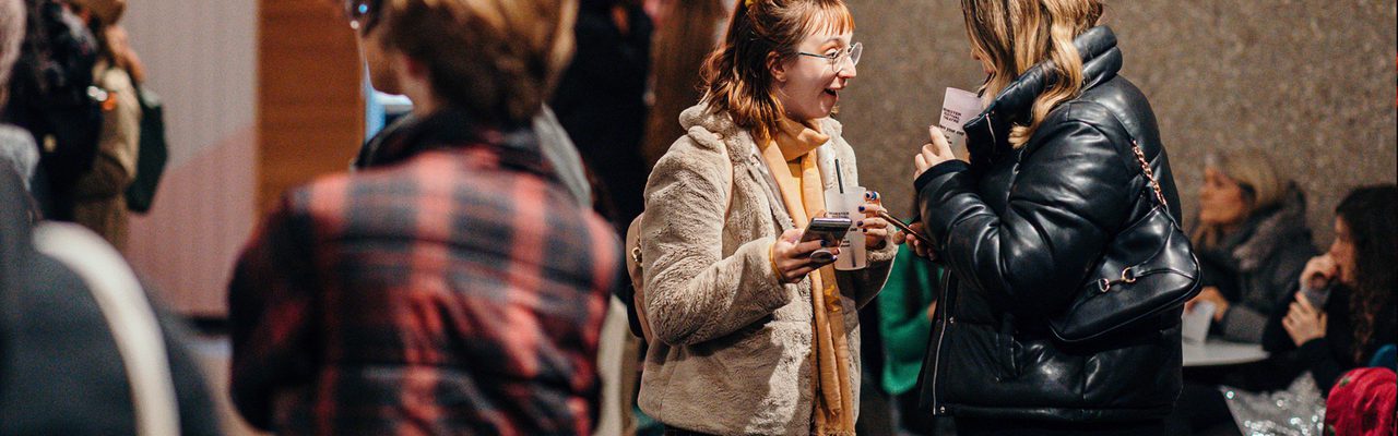 Two audience members stand in the Festival Theatre foyer. They are holing drinks and their phones. The person on the left is wearing a beige coat and has red hair. They excitedly smile at the person in front of them, whose face is obscured. Another group stands closer to the camera with their faces obscured.